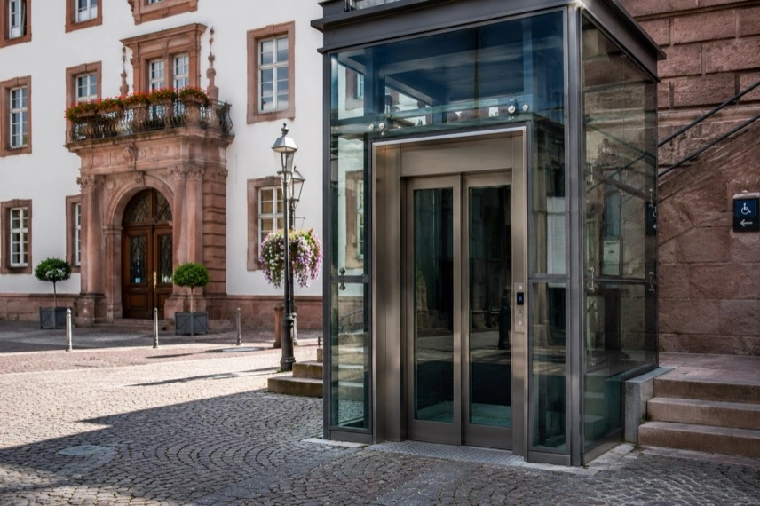 Moderne gläserne Aufzugskabine neben einem historischen Platz mit Natursteinpflaster und öffentlichem Gebäude im Hintergrund.