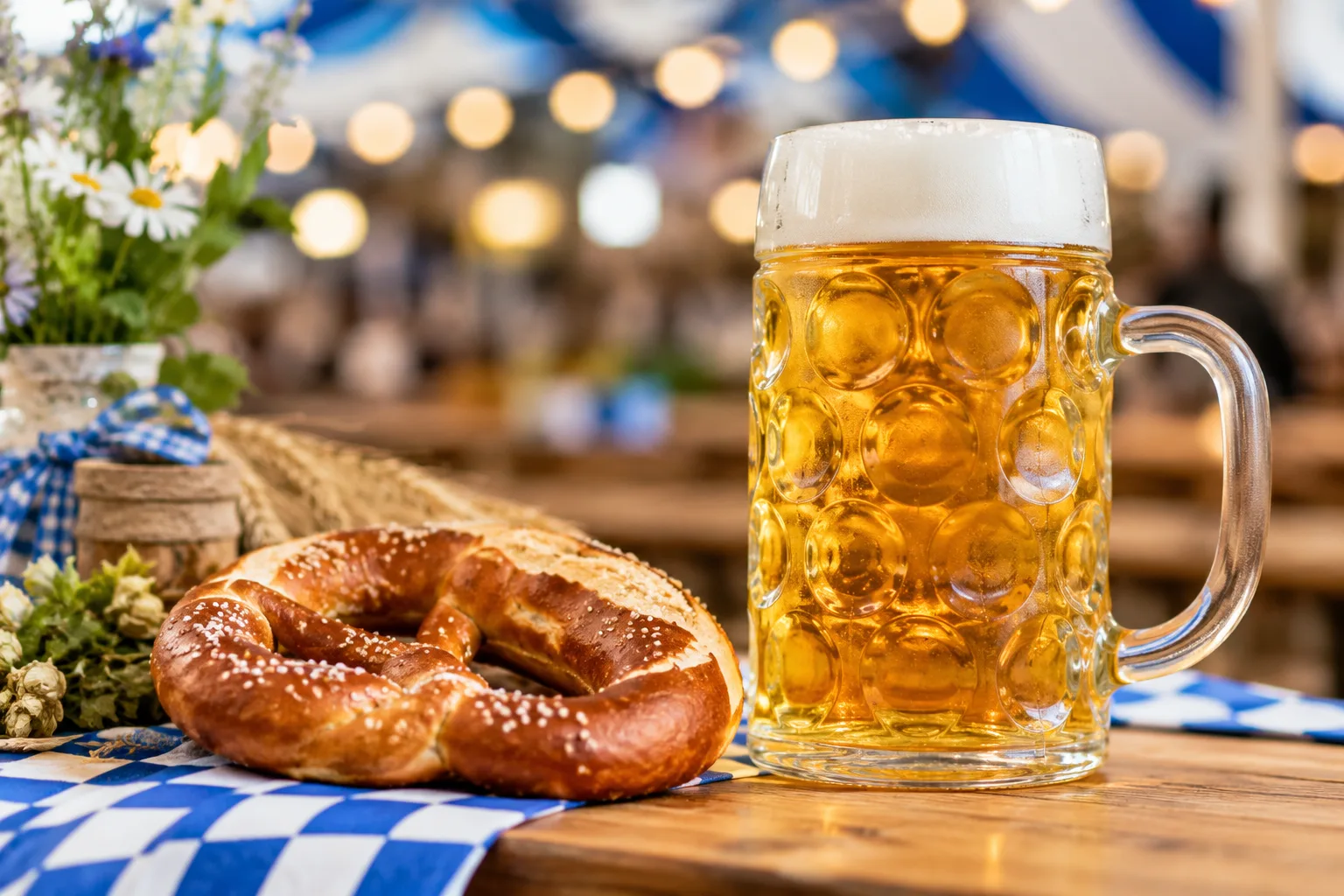 Bierkrug mit Brezel auf einem Holztisch mit blau-weißer Tischdecke als traditionelles Oktoberfest-Motiv.