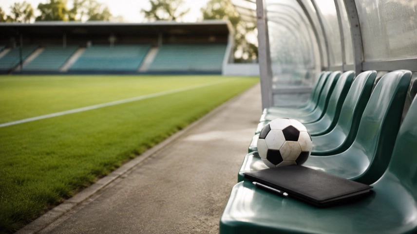 Leere Trainerbank mit Fußball und Taktikmappe an einem neutralen Fußballplatz mit Tribüne im Hintergrund.