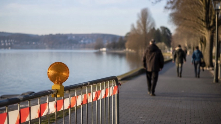 Öffentlicher Uferweg an einem See mit allgemeinem Sicherheitshinweis und anonymen Passanten im Hintergrund