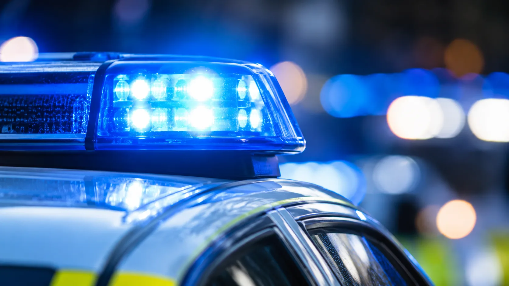 Blaues Polizeiblaulicht auf dem Dach eines neutralen Polizeifahrzeugs vor dunklem, unscharfem Hintergrund.
