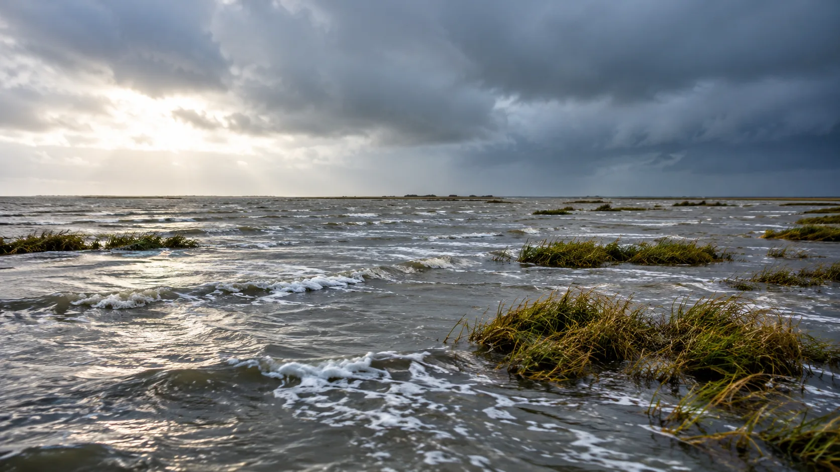 Überflutete Wattlandschaft an der Nordseeküste mit Grasbüscheln, erhöhtem Wasserstand und bewölktem Himmel.