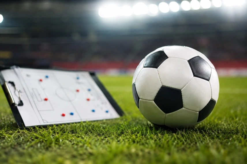 Fußball auf einem Rasen, im Hintergrund zwei anonyme Spieler in roten Trainingsoberteilen.