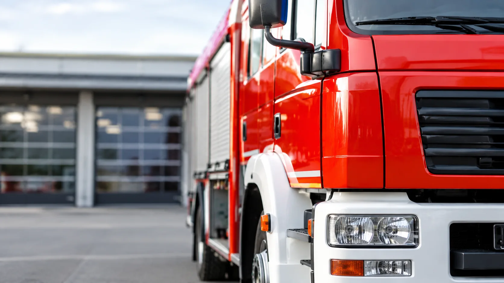 Rotes Feuerwehrfahrzeug mit Warnleuchte und seitlichen Ausrüstungsfächern auf einer freien Fläche.