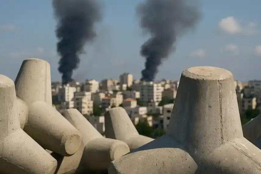 Betonbarrieren im Vordergrund mit einer Stadt im Hintergrund, aus der dunkle Rauchwolken aufsteigen, unter klarem Himmel.