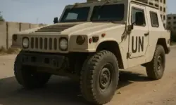 Beiges UN-Panzerfahrzeug auf einer staubigen Straße vor beschädigten Gebäuden bei Tageslicht.