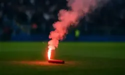 Rote Pyrofackel brennt auf grünem Fußballrasen, dichter Rauch steigt in die Luft, Hintergrund mit unscharfen Zuschauern.
