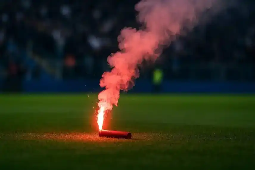 Rote Pyrofackel brennt auf grünem Fußballrasen, dichter Rauch steigt in die Luft, Hintergrund mit unscharfen Zuschauern.