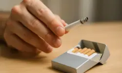 Nahaufnahme einer Hand, die eine Zigarette über einer geöffneten Schachtel mit Filterzigaretten hält, auf einem hellen Holztisch.