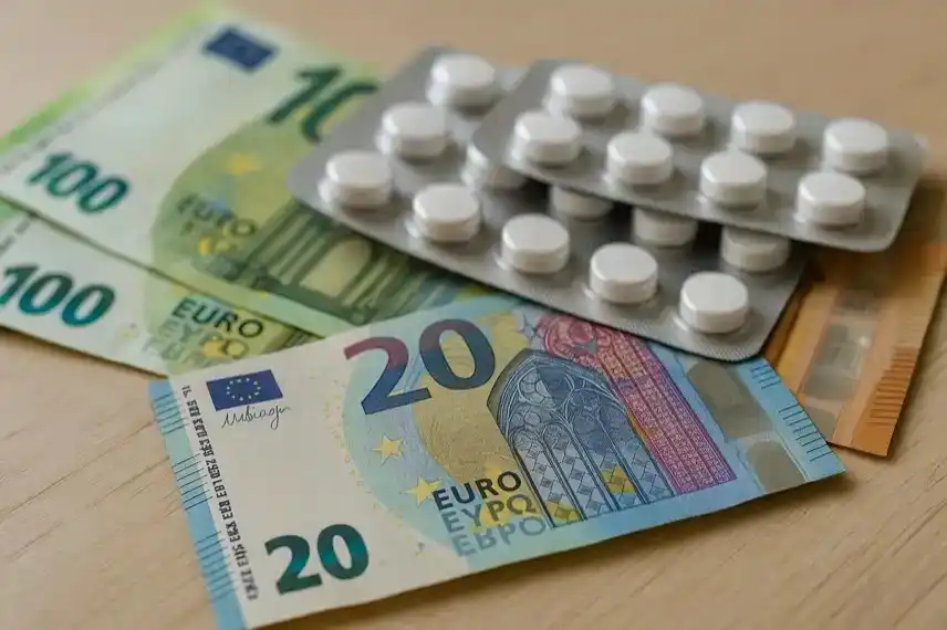 Euro-Banknoten und Blisterpackungen mit weißen Tabletten liegen auf einem hellen Holztisch.