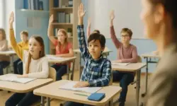 Mehrere Kinder sitzen in einem hellen Klassenzimmer an ihren Tischen und melden sich, während die Lehrerin vorne steht.