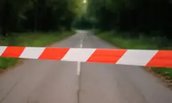 Rote und weiße Absperrband-Markierung quer über eine leere Landstraße im Wald mit weicher Tiefenschärfe im Hintergrund.