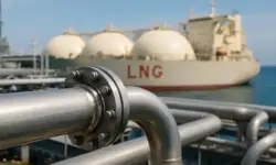 Nahaufnahme einer silbernen Gasleitung mit Schraubverbindung, im Hintergrund ein LNG-Tankschiff im Hafen bei Tageslicht.