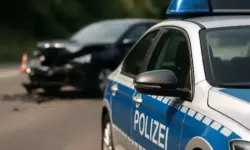 Polizeiauto mit blauer Aufschrift „Polizei“ im Vordergrund, im Hintergrund ein beschädigter schwarzer Pkw nach einem Unfall.