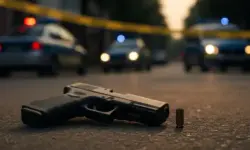 Eine Pistole und eine Patronenhülse liegen auf einer Straße, im Hintergrund sind unscharfe Polizeifahrzeuge mit Blaulicht zu sehen.