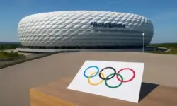 Die Allianz Arena in München bei Tageslicht, im Vordergrund ein Blatt Papier mit den fünf olympischen Ringen auf einem Tisch.