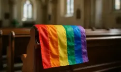 Regenbogenflagge liegt über der Lehne einer hölzernen Kirchenbank, mit unscharfem Kircheninneren im Hintergrund.