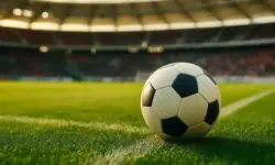 Ein Fußball liegt auf grünem Rasen in einem Stadion bei Sonnenlicht, unscharfer Hintergrund mit Tribünen und Zuschauern.