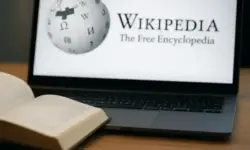 Ein geöffnetes Buch liegt auf einem Holztisch vor einem Laptop, dessen Bildschirm eine Online-Enzyklopädie zeigt.