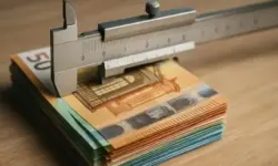 Nahaufnahme eines Messschiebers, der einen Stapel farbiger Eurobanknoten auf einem Holztisch misst.