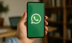 Hand hält ein Smartphone mit WhatsApp-Logo auf dem Display, im Hintergrund unscharf ein Schreibtisch mit Laptop und Regal.