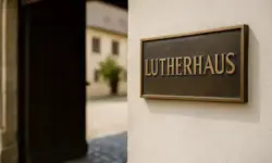 Bronzene Tafel mit der Aufschrift „Lutherhaus“ an einer hellen Hauswand neben einer geöffneten Holztür, im Hintergrund ein unscharfer Innenhof.