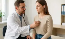 Ein Arzt hört mit einem Stethoskop den Brustbereich einer jungen Frau in einer hellen Arztpraxis ab.