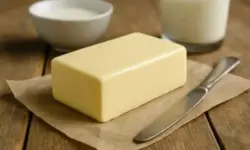 Ein Stück Butter liegt auf braunem Papier neben einem Messer, im Hintergrund sind Milch und eine Schale zu sehen.