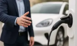 Ein Mann steht neben einer weißen Elektroauto-Ladestation und greift nach dem Ladekabel, im Hintergrund ein weißes Auto.