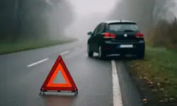 Ein Auto steht am Straßenrand einer nebligen Landstraße, im Vordergrund steht ein aufgestelltes rotes Warndreieck auf nasser Fahrbahn.