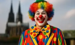 Ein Clown mit farbenfrohem Kostüm und Perücke steht draußen, im Hintergrund unscharf der Kölner Dom bei Tageslicht.