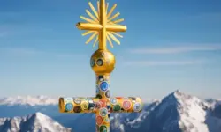Ein goldenes Gipfelkreuz mit bunten Stickern vor schneebedeckten Bergen und blauem Himmel.