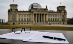Dokumente, Brille und Stift auf einem Tisch mit unscharfem Reichstagsgebäude im Hintergrund.