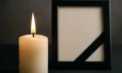 Brennende helle Kerze im Vordergrund, daneben ein unscharfer schwarzer Rahmen mit diagonalem Trauerband.