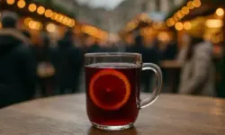 Ein klarer Glasmug mit heißem Glühwein und Orangenscheibe steht auf einem Holztisch, im Hintergrund unscharfe Lichter eines Weihnachtsmarktes.