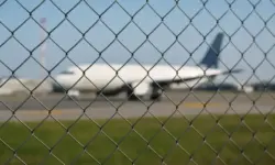 Symbolisches Foto eines Flughafenvorfelds mit unscharfem Flugzeug und Zaun bei Tageslicht.