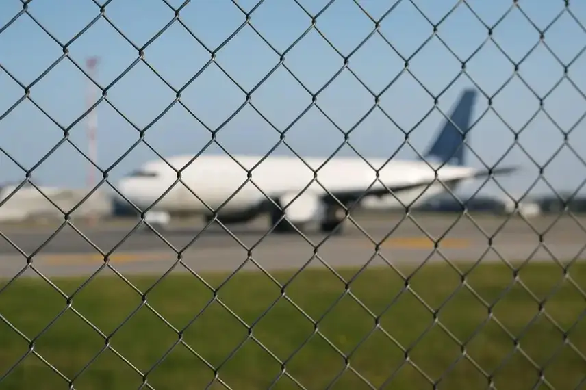 Symbolisches Foto eines Flughafenvorfelds mit unscharfem Flugzeug und Zaun bei Tageslicht.