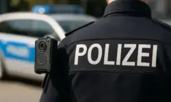 Rückansicht eines deutschen Polizisten mit Bodycam auf der Schulter, im Hintergrund ein unscharfes Polizeifahrzeug.