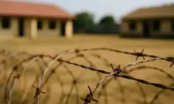 Stacheldraht im Vordergrund mit unscharfen Schulgebäuden im Hintergrund auf trockenem Gelände.