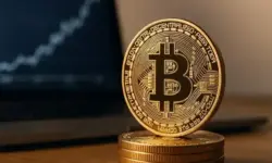 Goldene Bitcoin-Münze auf einem Holztisch vor einem Laptop mit unscharfem Kursdiagramm im Hintergrund.