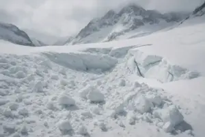 Verschneite Alpenlandschaft mit Schneebruch, Felsen und Berggipfeln unter bewölktem Himmel.