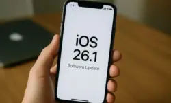 Eine Hand hält ein iPhone mit der Anzeige „iOS 26.1 Software Update“ vor einem unscharfen Hintergrund mit einem Laptop.