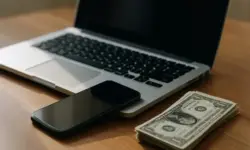 Laptop, Smartphone und ein Stapel Geldscheine auf einem Holztisch bei natürlichem Licht.