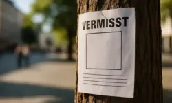 Nahaufnahme eines weißen Vermisstenzettels ohne Foto, befestigt an der Rinde eines Baumes, mit verschwommenem Stadtplatz im Hintergrund.