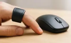 Ein Finger trägt einen schwarzen Smart-Ring und liegt neben einer schwarzen Computermaus auf einem hellen Holztisch.
