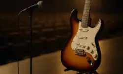 Eine E-Gitarre steht neben einem Mikrofonständer auf einer leeren Konzertbühne, im Hintergrund unscharfe Zuschauerreihen.