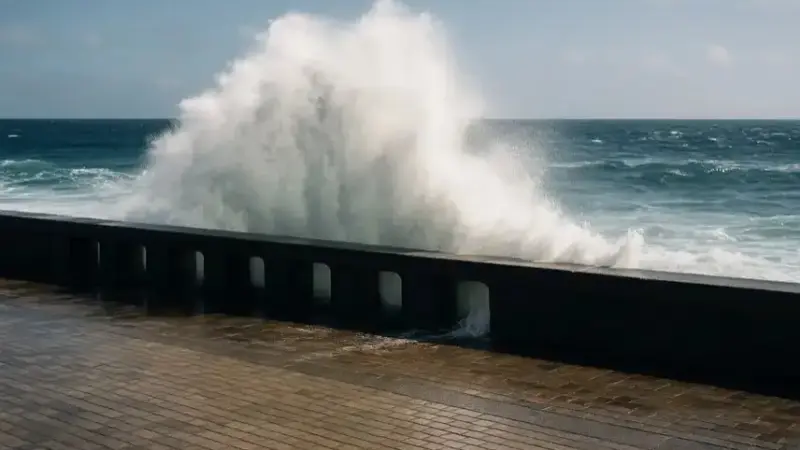 Hohe Welle bricht über eine Meeresmauer an einer sonnigen Küstenpromenade.