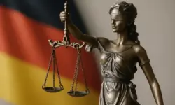 Bronzestatue der Justitia mit Waage vor unscharfer deutscher Flagge im Hintergrund.