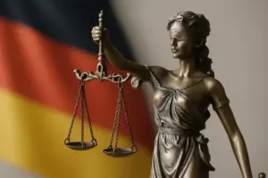 Bronzestatue der Justitia mit Waage vor unscharfer deutscher Flagge im Hintergrund.