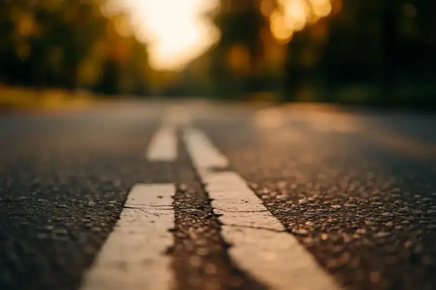 Asphaltstraße mit Mittelstreifen, aufgenommen aus Bodennähe bei warmem Sonnenlicht, Hintergrund unscharf mit Bäumen.